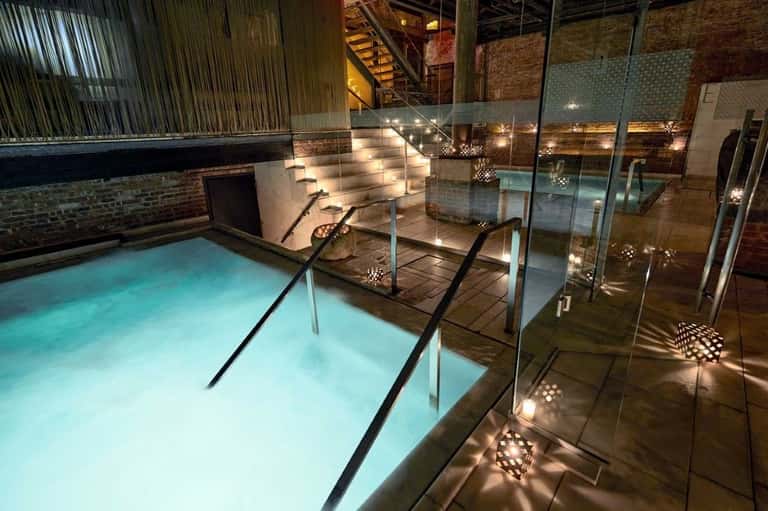Indoor thermal pool with turquoise water, glass railings, and ambient lighting in a brick industrial space