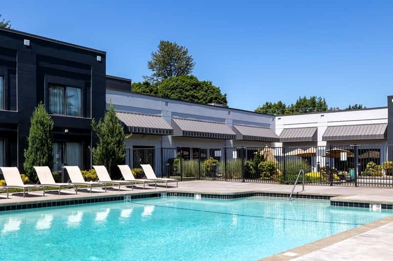 Resort swimming pool with lounge chairs, modern black and white buildings, metal fence, and trees under clear blue sky