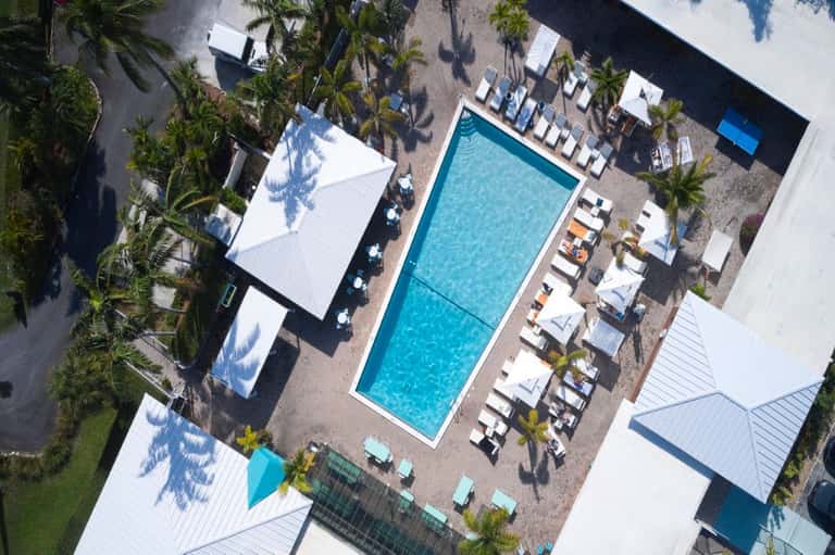 Aerial view of resort pool with white loungers, palm trees, and white-roofed buildings surrounding the pool deck