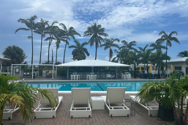 Resort pool with white loungers, large white tent pavilion, palm trees, and tropical landscaping under blue sky