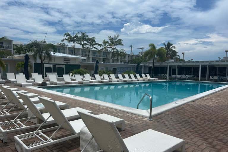 Resort swimming pool with turquoise water, white lounge chairs, palm trees, and multi-story hotel building under blue sky
