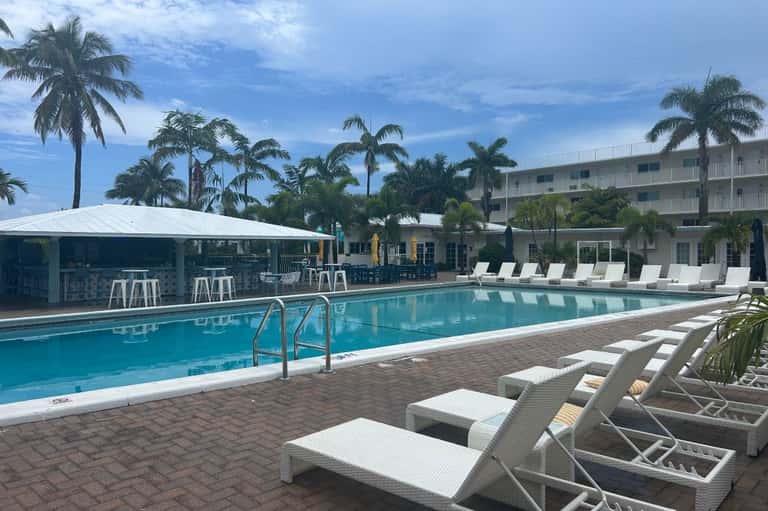 Pool area with white lounge chairs, bar pavilion, palm trees, and residential buildings in tropical resort setting