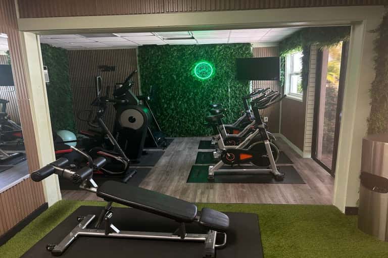 Modern fitness center with stationary bikes, exercise equipment, green accent wall with neon clock, and artificial turf flooring