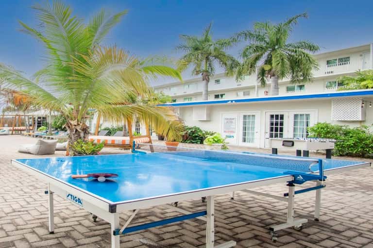 Outdoor ping pong table on brick patio with palm trees, resort buildings, and clear sky in background