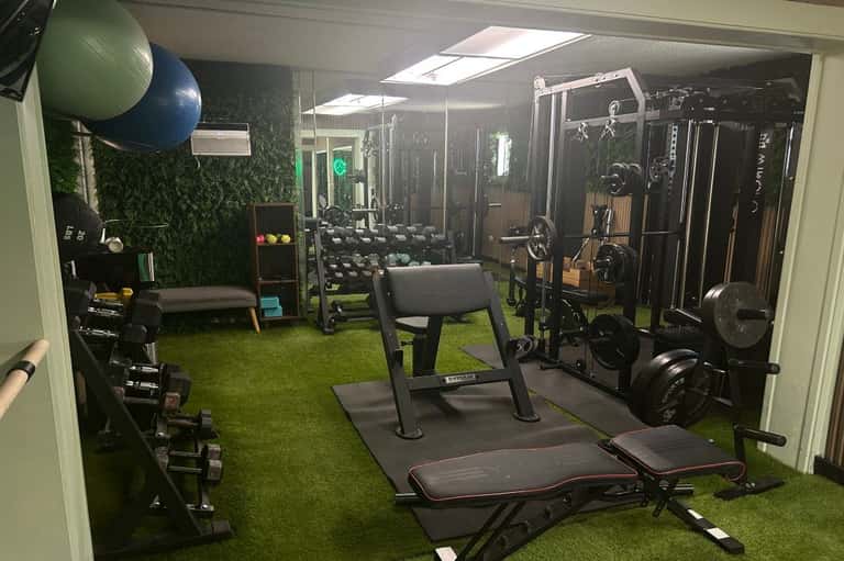 Fully equipped gym with cardio machines, weight stations, dumbbells, green wall accent, and artificial turf floor