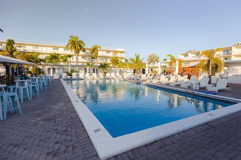 Resort pool with white lounge chairs, blue umbrellas, palm trees, and multi-story residential buildings at dusk
