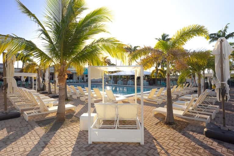 Resort pool with white lounge chairs, palm tree shade structures, pergolas, and calm blue water surrounded by tropical vegetation