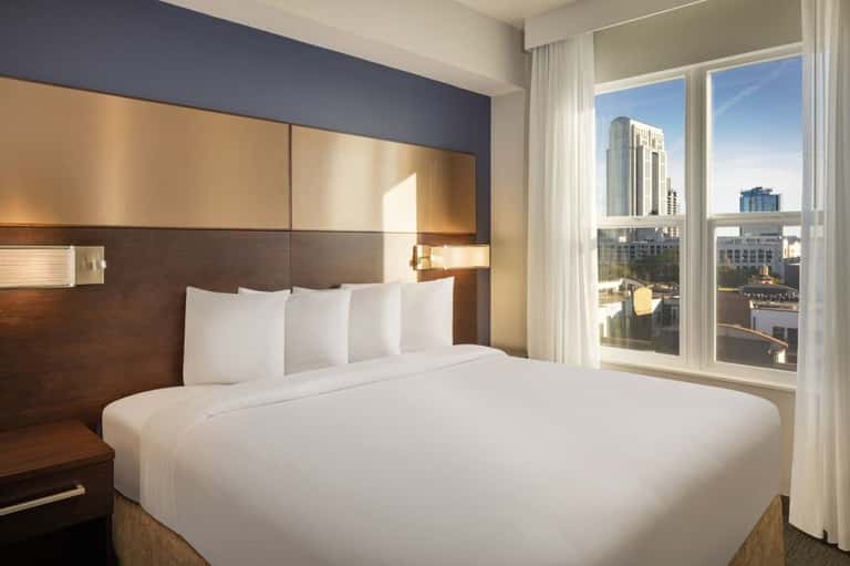 Modern hotel bedroom with king bed, dark headboard, city skyline views through floor-to-ceiling windows