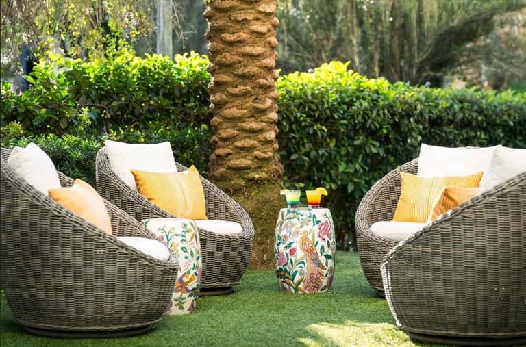 Two wicker lounge chairs with white and yellow pillows on grass beside palm tree and tropical greenery