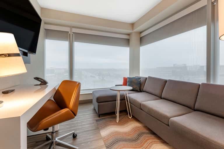 Modern hotel suite with floor-to-ceiling windows, tan sofa, orange leather desk chair, and cityscape views