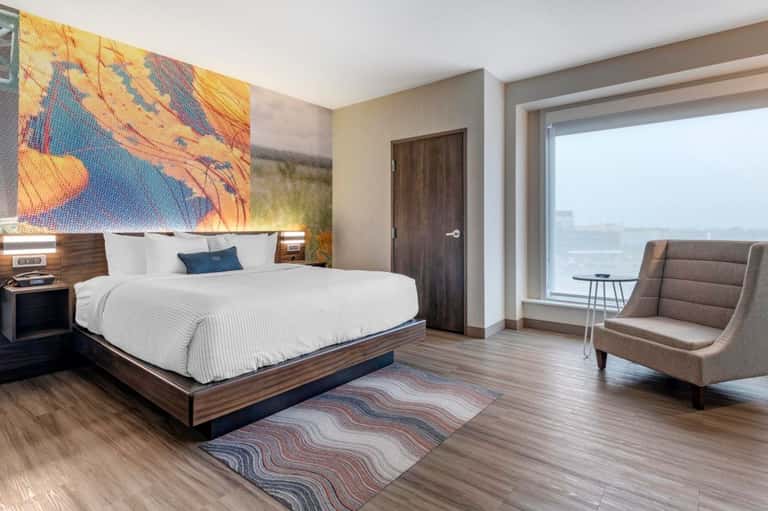 Modern hotel bedroom with king bed, colorful headboard art, sitting area, and expansive window views