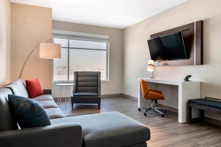 Hotel suite living area with sofa, desk with orange chair, flat-screen TV, and city views through windows