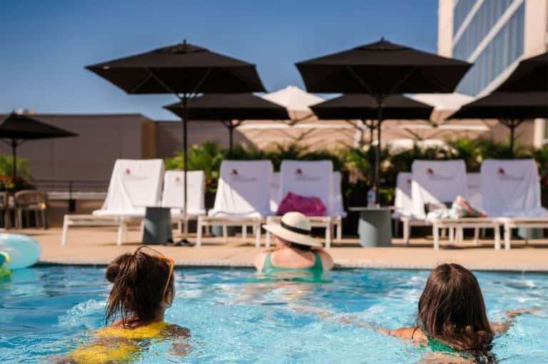 Two children swimming in a bright blue pool with black umbrellas and white lounge chairs visible in the background