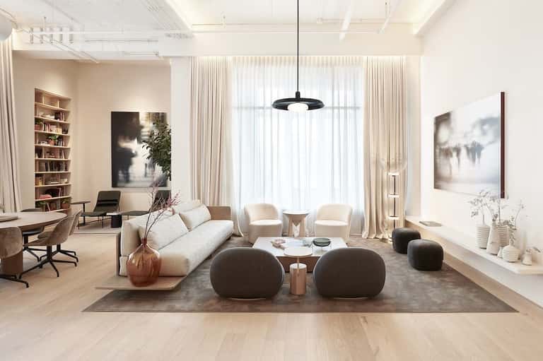 Sophisticated living room with cream sofa, round ottomans, bookshelf, modern pendant lighting, and expansive curtained windows