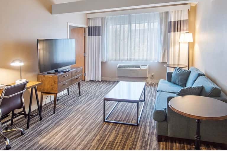 Spacious hotel suite with living area, television, blue sofa, striped carpet, and floor-to-ceiling windows