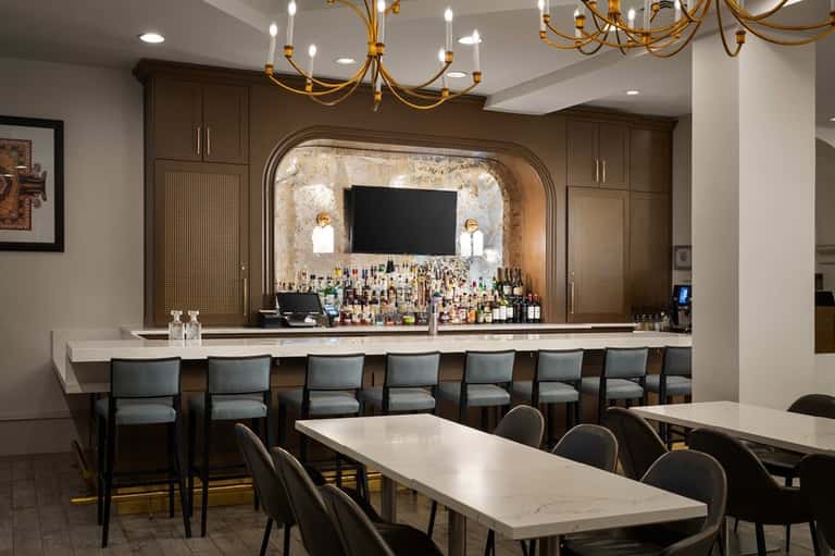 Elegant hotel bar with brown cabinetry, gold chandelier, white marble counter, and gray upholstered bar seating