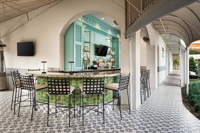 Outdoor bar with turquoise accent wall, black checkered chairs, patterned tile flooring under arched colonnade