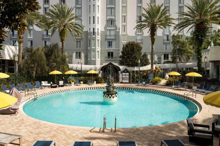 Resort pool surrounded by palm trees, yellow umbrellas, lounge chairs, and multi-story hotel building