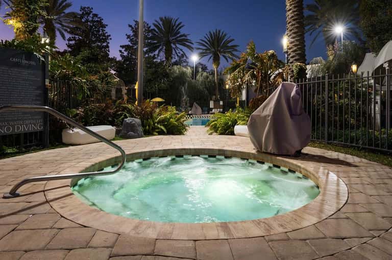 Illuminated spa hot tub at night with palm trees, landscaping, and covered lounge chairs nearby