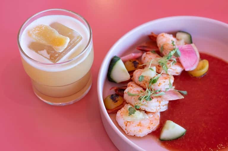 Cocktail with grapefruit garnish and plated shrimp appetizer on pink surface