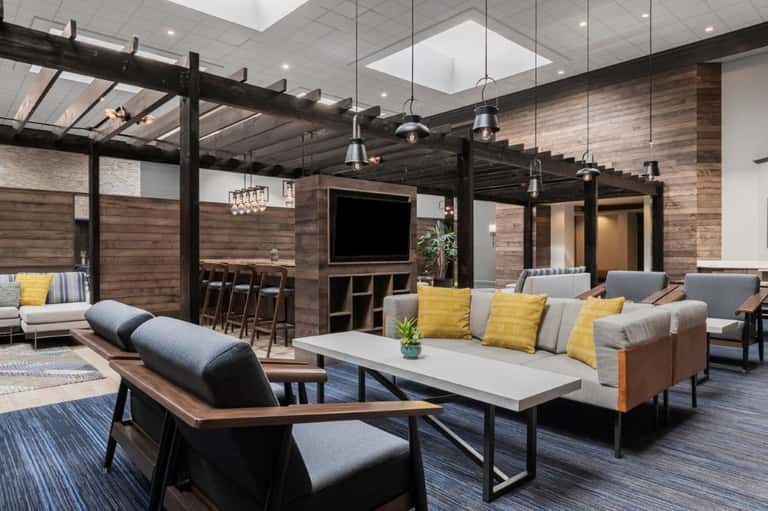 Contemporary lounge area with wood-paneled walls, black pergola, hanging pendant lights, gray seating, and yellow accent pillows