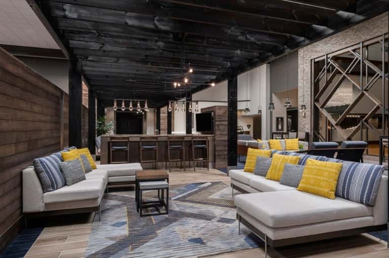 Upscale bar lounge with dark wood bar, black ceiling beams, cream sectional seating, patterned throw pillows, and decorative lighting