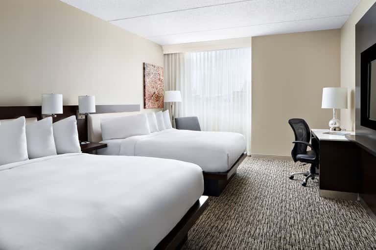 Hotel guest room with two queen beds, dark wood headboards, cream walls, work desk, and window with sheer curtains