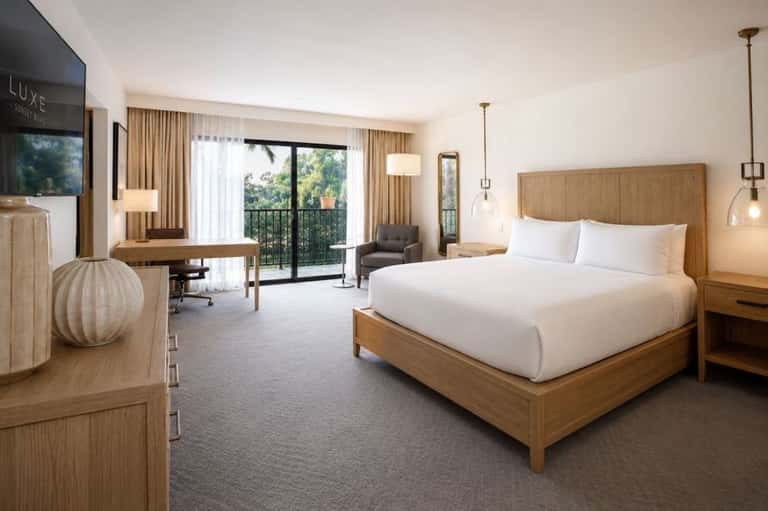 Luxury hotel bedroom with wooden bed, white linens, desk, and balcony access to greenery