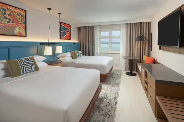 Hotel room with two queen beds, blue paneled headboard, pendant lighting, and water views