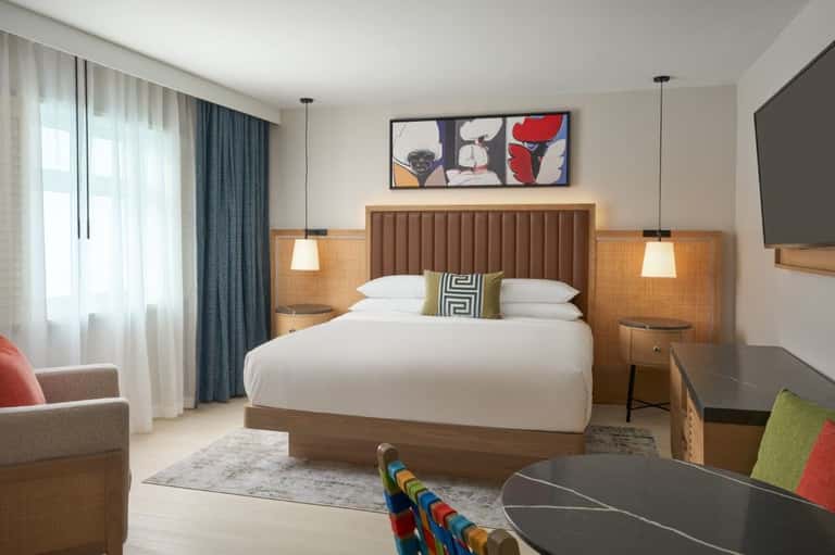 Contemporary hotel bedroom with wood-framed bed, pendant lighting, flat-screen TV, and seating area