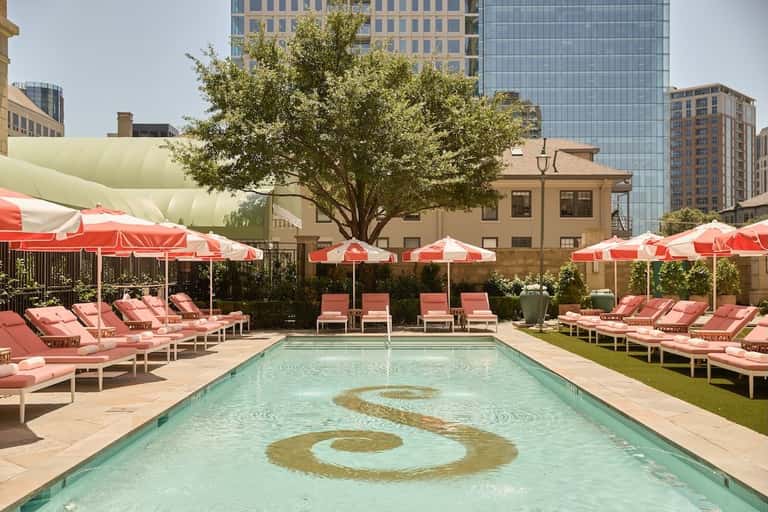 Urban rooftop pool with coral pink loungers under red umbrellas, surrounded by city skyscrapers and trees