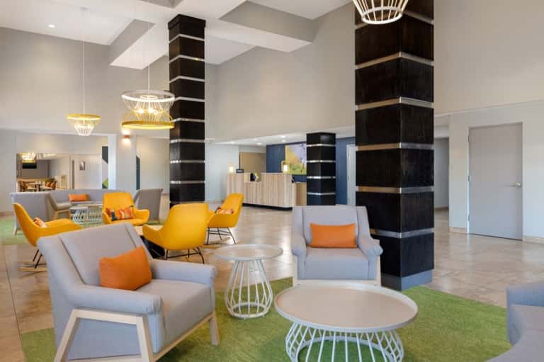 Modern hotel lobby with gray and yellow chairs, pendant lighting, dark wood pillars, and green area rugs