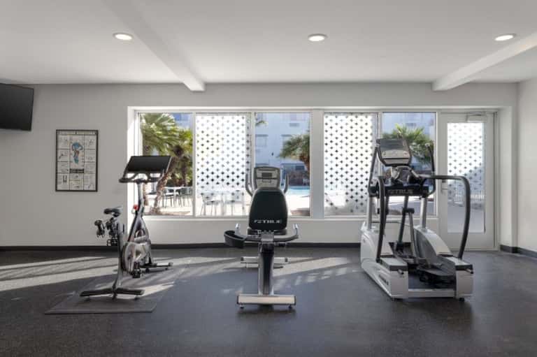 Bright fitness room with stationary bikes and elliptical equipment, overlooking pool through glass doors