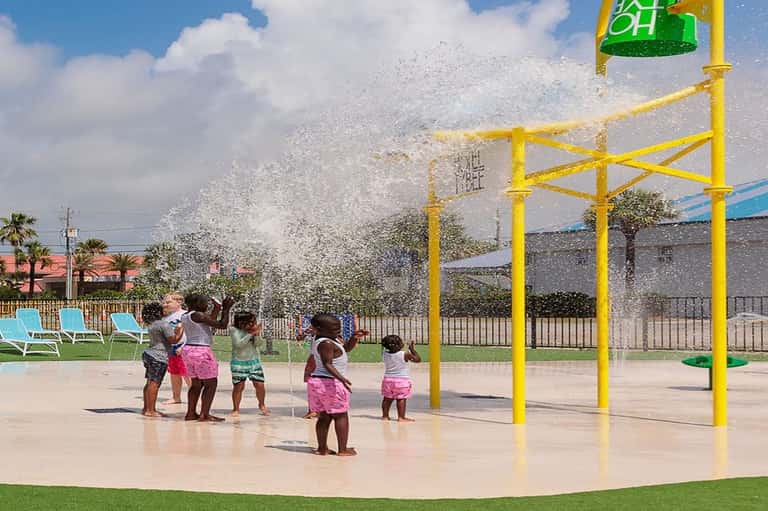 Children playing under yellow water spray structure with bucket dump feature on wet deck