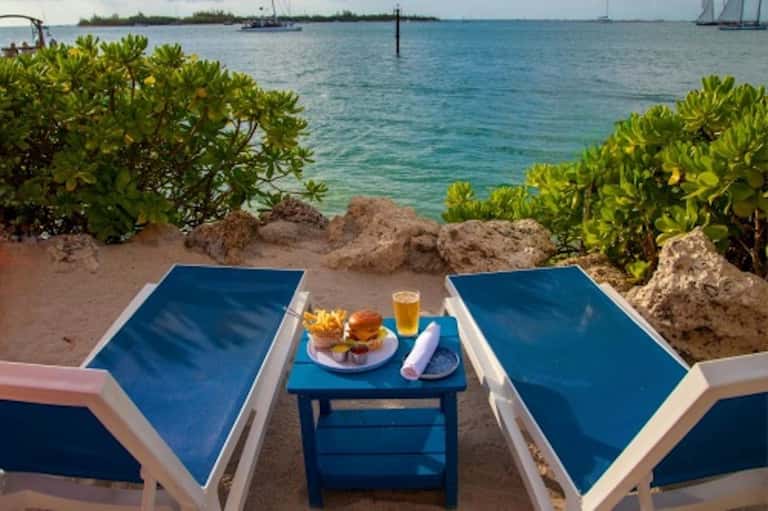 Beach loungers with food and beverage on sand surrounded by mangroves and turquoise ocean