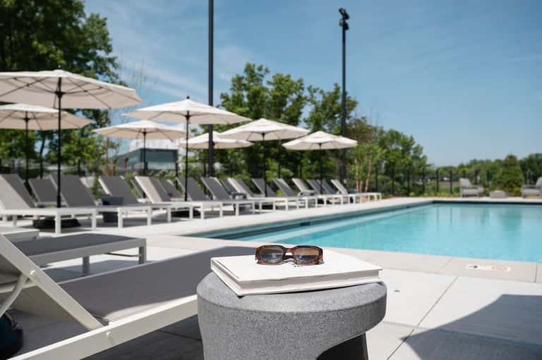 Modern resort pool deck with white umbrellas, gray loungers, and manicured landscaping under blue sky