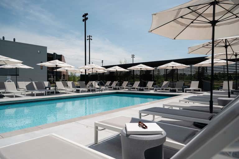 Contemporary pool area with white umbrellas, gray lounge chairs, and black privacy fence alongside building