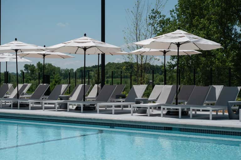 Resort pool with white umbrellas, gray lounge chairs, black poles, and lush green treeline backdrop