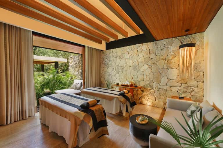 Spa treatment room with twin massage beds, stone accent wall, wooden ceiling beams, and garden views