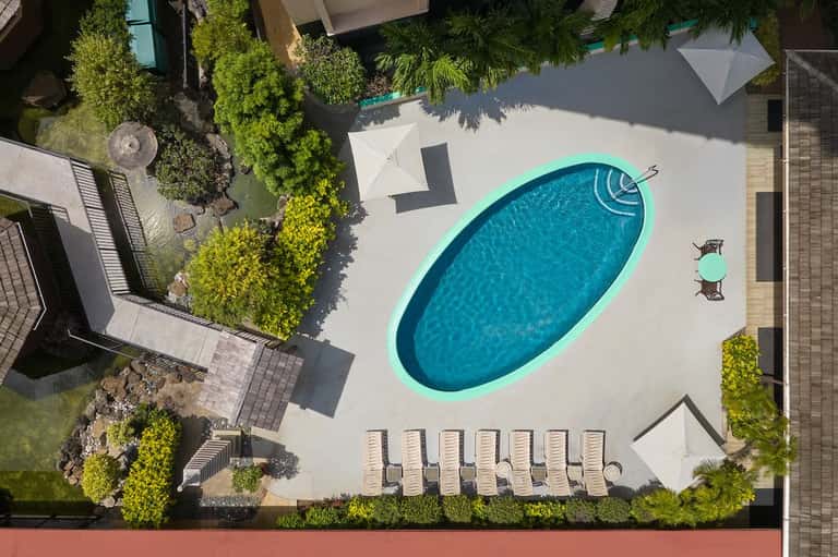 Aerial view of oval-shaped pool with turquoise deck trim, white umbrellas, lounge chairs, and landscaped courtyard