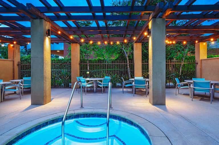 Covered poolside lounge area with spa tub, turquoise seating, warm lighting, and garden views