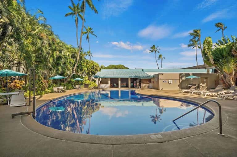 Resort pool surrounded by tall palm trees, lounge chairs, turquoise umbrellas, and ocean view in background