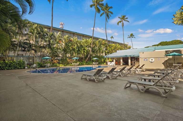 Pool area with turquoise deck trim, lounge chairs, palm trees, multi-story hotel building, and blue sky