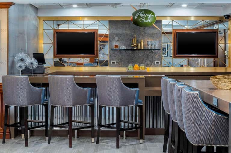 Hotel bar with dark marble counter, bar stools, dual TVs, modern lighting, and green sign