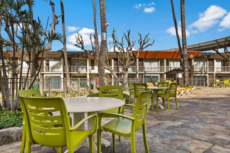 Outdoor patio area with bright lime-green chairs, white round tables, palm trees, and motel buildings in background.