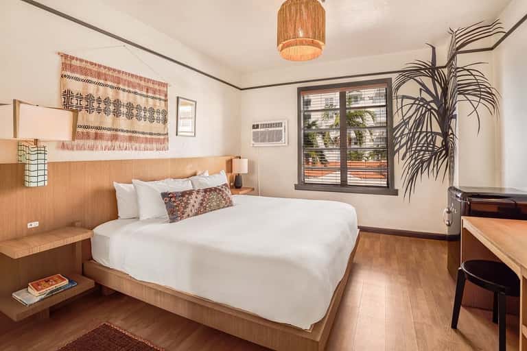 Modern hotel bedroom with wooden bed frame, white linens, woven wall hanging, potted plant, and large windows with views
