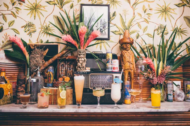 Tropical-themed bar with floral wallpaper, wooden carved figures, pink flowers, vintage radio, and colorful cocktails on counter