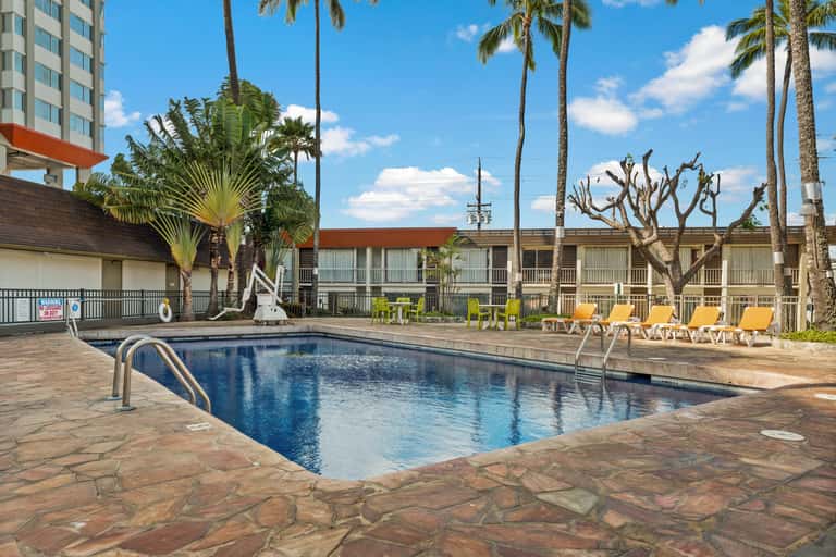 Resort swimming pool with blue water, lounge chairs, palm trees, and multi-story hotel buildings with orange accents.