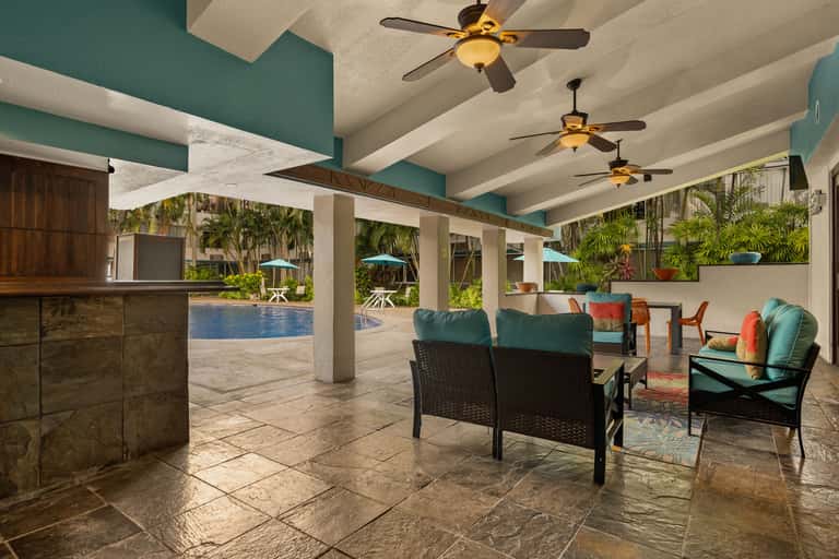 Covered resort patio with lounge seating, ceiling fans, and swimming pool visible beyond