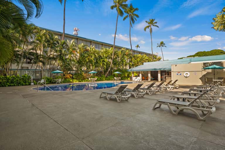Resort swimming pool surrounded by lounge chairs, palm trees, and hotel building